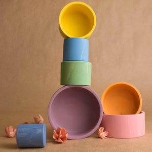 May include: A collection of brightly coloured ceramic bowls in a still-life arrangement. The bowls are stacked in a tower, with colours including yellow, blue, green, and purple. Peach-coloured floral decorations are scattered around.
