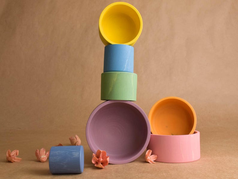 May include: A collection of brightly coloured ceramic bowls in a still-life arrangement. The bowls are stacked in a tower, with colours including yellow, blue, green, and purple. Peach-coloured floral decorations are scattered around.