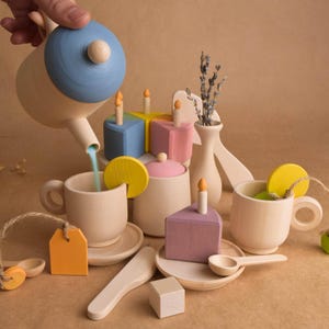May include: Wooden toy tea set with a teapot pouring into a teacup. The set includes a sugar bowl, cake slices, spoons, and a vase with dried flowers. The teapot has a blue lid, and the cake slices are painted in pastel colors.