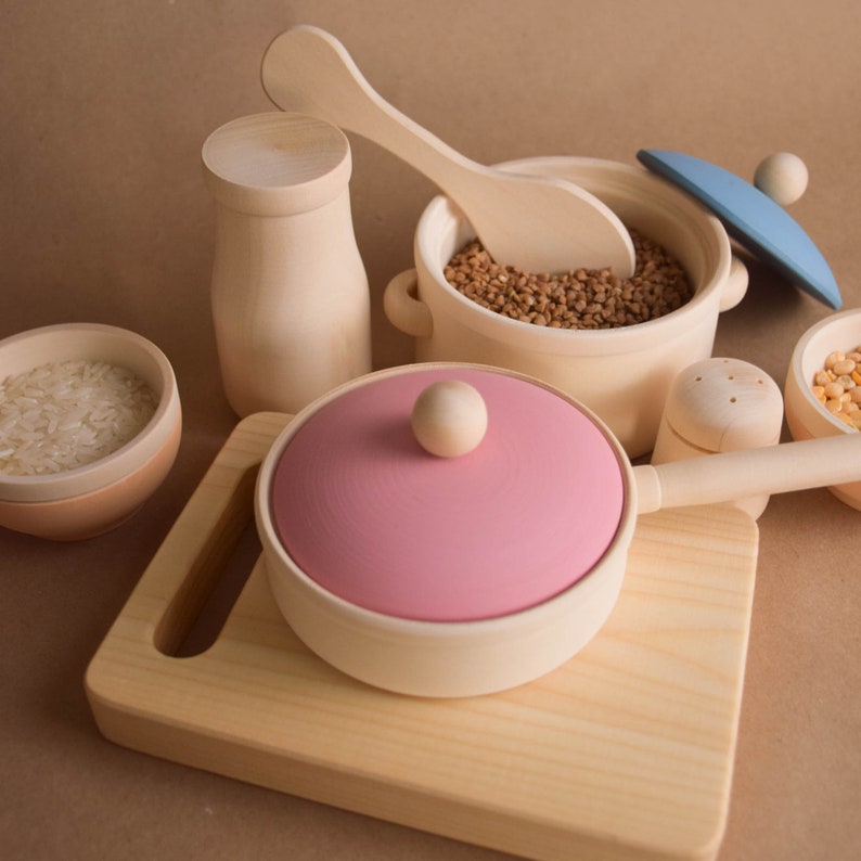May include: A set of wooden play kitchen utensils, including a pink pot with a lid, a wooden spoon, a wooden salt shaker, a wooden container, a bowl of rice, and a pot filled with brown grains.