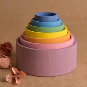 May include: A wooden rainbow stacking toy with seven colourful, graduated rings. The colours include purple, pink, orange, yellow, green, blue, and a darker blue. The toy is on a brown surface with dried flowers.