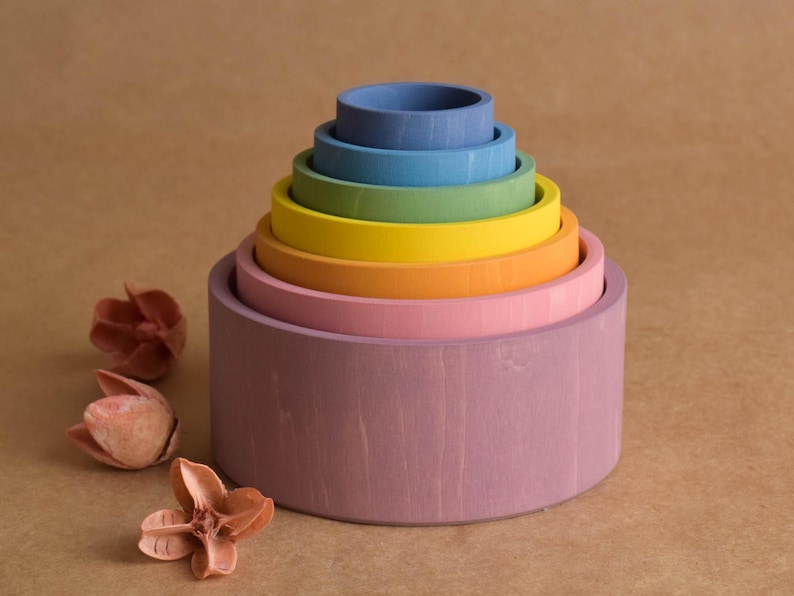 May include: A wooden rainbow stacking toy with seven colourful, graduated rings. The colours include purple, pink, orange, yellow, green, blue, and a darker blue. The toy is on a brown surface with dried flowers.