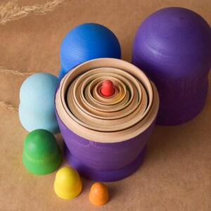Pode incluir: Um conjunto de bonecas russas de madeira coloridas, ou bonecas empilh&aacute;veis, com uma base roxa e um arco-&iacute;ris de cores no centro. As bonecas est&atilde;o dispostas em c&iacute;rculo com a maior boneca no centro.