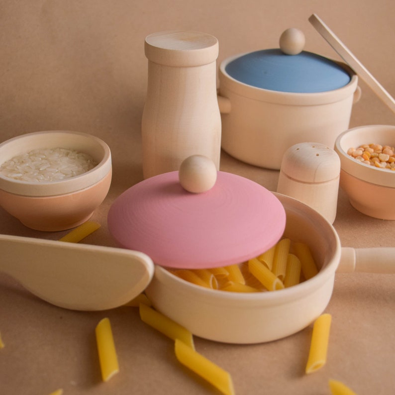 May include: A set of wooden toy kitchen cookware, including a pink pot with lid, a blue pot with lid, a small bowl with rice, a salt shaker, and a milk jug. The cookware is arranged on a brown surface with pasta scattered around.