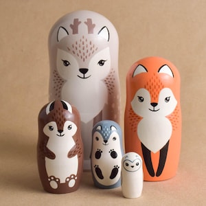 May include: A set of five wooden nesting dolls featuring stylized animal illustrations. The largest is a beige deer, followed by an orange fox, a brown squirrel, a blue bear, and a small white owl, all with painted faces and markings.