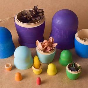 Pode incluir: Um conjunto de bonecas russas de madeira coloridas, ou bonecas matrioska, com v&aacute;rios desenhos. As bonecas est&atilde;o dispostas em c&iacute;rculo sobre uma superf&iacute;cie marrom. A boneca maior &eacute; roxa e tem um cone de pinho dentro. A boneca menor &eacute; amarela e tem uma pequena flor dentro.