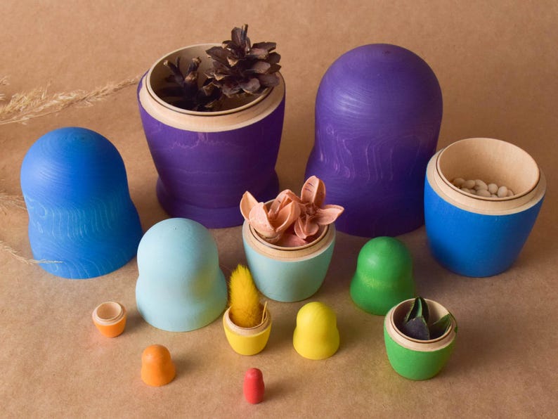 Pode incluir: Um conjunto de bonecas russas de madeira coloridas, ou bonecas matrioska, com v&aacute;rios desenhos. As bonecas est&atilde;o dispostas em c&iacute;rculo sobre uma superf&iacute;cie marrom. A boneca maior &eacute; roxa e tem um cone de pinho dentro. A boneca menor &eacute; amarela e tem uma pequena flor dentro.