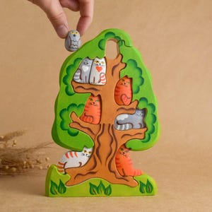 Könnte beinhalten: Ein Holzpuzzle in Form eines Baumes mit Katzen und einer Eule. Der Baum ist grün mit einem braunen Stamm, die Katzen sind orange, grau und weiß. Eine Hand hält die Eule über dem Baum.
