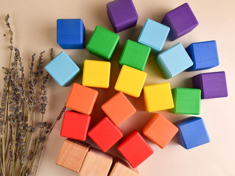 May include: A set of colorful wooden blocks in a rainbow pattern. The blocks are arranged in a circle on a beige surface. There is a bunch of dried lavender in the lower left corner of the image.