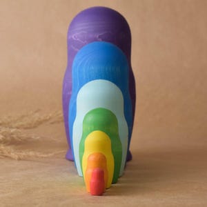 Pode incluir: Um conjunto de bonecas russas aninhadas com cores do arco-&iacute;ris. A maior boneca &eacute; roxa, seguida de azul, azul claro, verde, amarelo, laranja e vermelho.