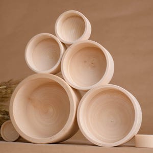 May include: A collection of unfinished wooden bowls of varying sizes, stacked in a pyramid shape. The bowls are light-colored with visible wood grain. A small, cylindrical wooden piece sits nearby. The background is a solid, neutral brown.