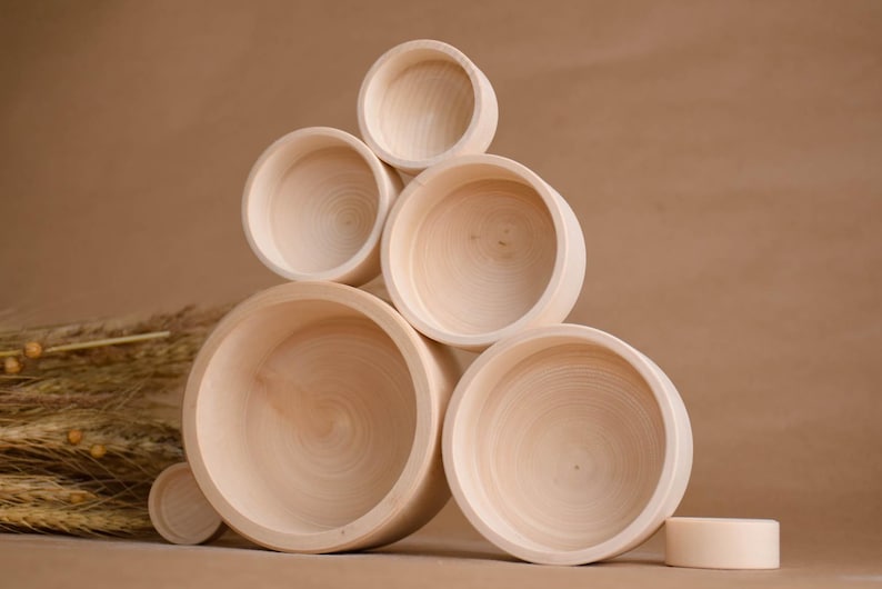 May include: A collection of unfinished wooden bowls of varying sizes, stacked in a pyramid shape. The bowls are light-colored with visible wood grain. A small, cylindrical wooden piece sits nearby. The background is a solid, neutral brown.