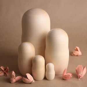May include: A set of unpainted wooden nesting dolls of varying sizes, displayed on a neutral backdrop. The dolls are smooth and show the natural wood grain. Several dried, pink floral decorations are scattered around the dolls.