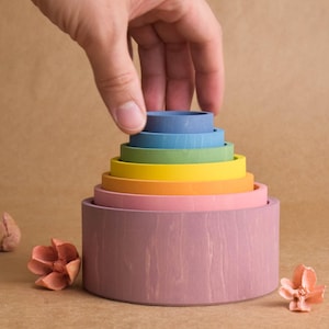 May include: A multi-coloured wooden stacking toy with a hand placing the top ring. The toy features rings in various colours, including pink, orange, yellow, green, blue, and purple. The toy is designed for children's play and learning.