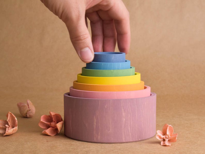 May include: A multi-coloured wooden stacking toy with a hand placing the top ring. The toy features rings in various colours, including pink, orange, yellow, green, blue, and purple. The toy is designed for children's play and learning.