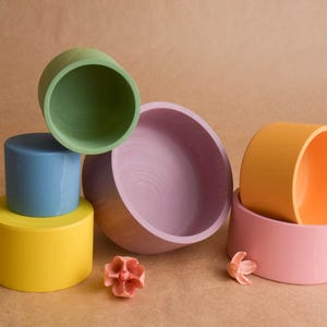 May include: A collection of brightly coloured wooden bowls and cylinders in a still life arrangement. The colours include yellow, blue, green, pink, and orange. Decorative floral elements are also present, creating a playful display.