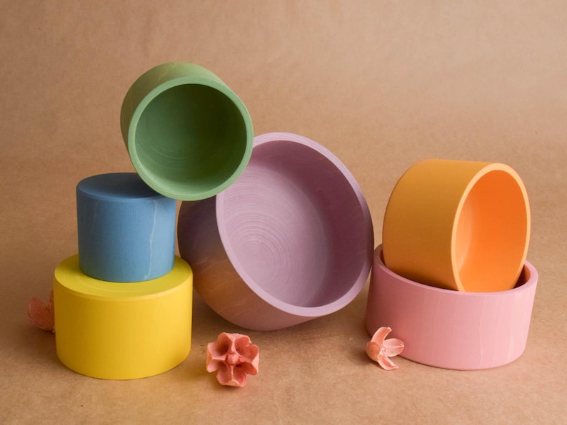 May include: A collection of brightly coloured wooden bowls and cylinders in a still life arrangement. The colours include yellow, blue, green, pink, and orange. Decorative floral elements are also present, creating a playful display.