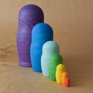 Pode incluir: Um conjunto de sete bonecas russas, tamb&eacute;m conhecidas como bonecas matrioska, pintadas em um esquema de cores arco-&iacute;ris. A boneca maior &eacute; roxa, seguida de azul, azul claro, verde, amarelo, laranja e vermelho.