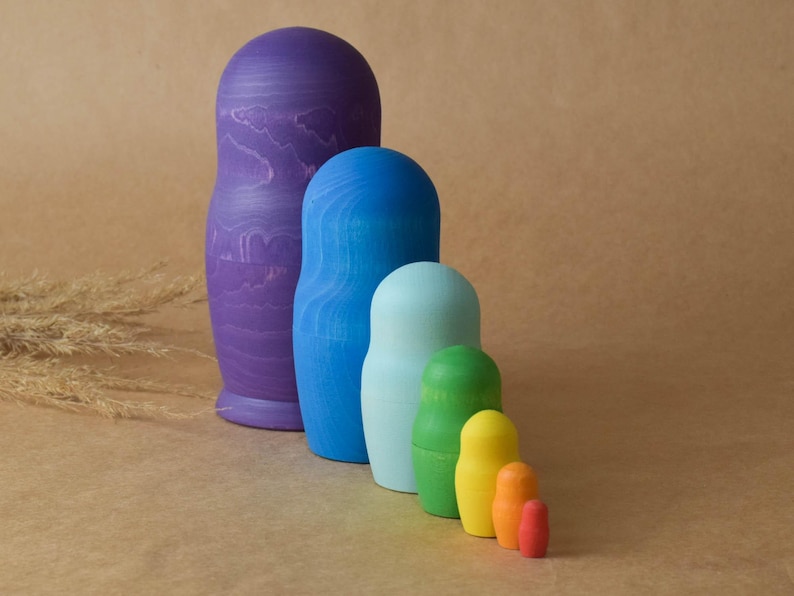 Pode incluir: Um conjunto de sete bonecas russas, tamb&eacute;m conhecidas como bonecas matrioska, pintadas em um esquema de cores arco-&iacute;ris. A boneca maior &eacute; roxa, seguida de azul, azul claro, verde, amarelo, laranja e vermelho.