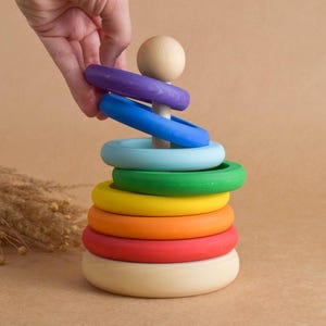 May include: A wooden stacking toy with a natural wood base and a series of colorful rings in red, orange, yellow, green, blue, and purple. A hand is placing a purple ring on the wooden peg. The toy is designed for young children.