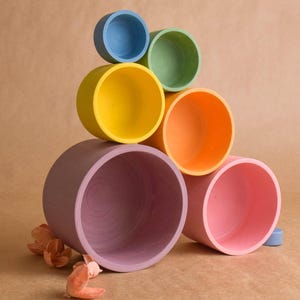 May include: A set of six colourful wooden nesting cups. The cups are stacked in a pyramid shape, with the largest cup at the bottom and the smallest cup at the top. The cups are painted in shades of purple, pink, orange, green, yellow, and blue.