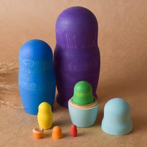 Pode incluir: Um conjunto de bonecas russas de madeira coloridas, ou bonecas de encaixe, de v&aacute;rios tamanhos. A boneca maior &eacute; roxa, a seguinte maior &eacute; azul e a menor &eacute; verde. Todas as bonecas s&atilde;o pintadas com desenhos simples.