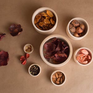 May include: Overhead shot of small, off-white bowls filled with various dried items, including petals, nuts, and peppercorns. The bowls are arranged on a brown surface, with some items scattered around them.
