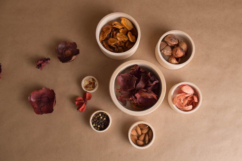 May include: Overhead shot of small, off-white bowls filled with various dried items, including petals, nuts, and peppercorns. The bowls are arranged on a brown surface, with some items scattered around them.