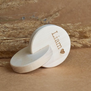 May include: A light-colored wooden baby name announcement. The name "Liam" is engraved on the top piece, accompanied by a small heart. The announcement is made of two interlocking wooden pieces.