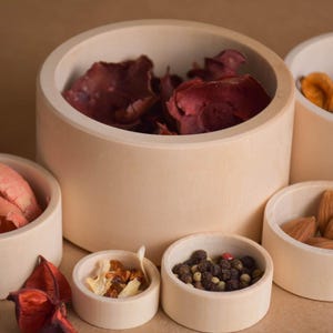 May include: A collection of light-colored wooden bowls of various sizes, filled with dried botanicals and spices. The bowls contain items like dried petals, almonds, and peppercorns. The arrangement is set against a neutral background.
