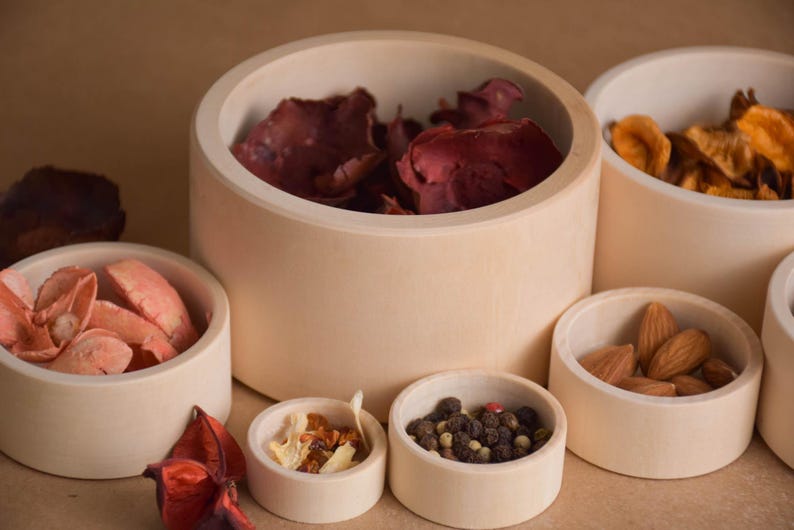 May include: A collection of light-colored wooden bowls of various sizes, filled with dried botanicals and spices. The bowls contain items like dried petals, almonds, and peppercorns. The arrangement is set against a neutral background.