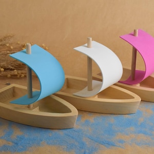 May include: Three wooden toy boats with coloured sails. One has a light blue sail, one has a white sail, and the third has a pink sail. The boats are set on a surface with blue and tan accents.