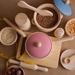 May include: A set of wooden play kitchen accessories, including a pink pot with a lid, a wooden spoon, a bowl of rice, a bowl of brown grains, a small wooden salt shaker, a bowl of yellow grains, and a wooden rolling pin.