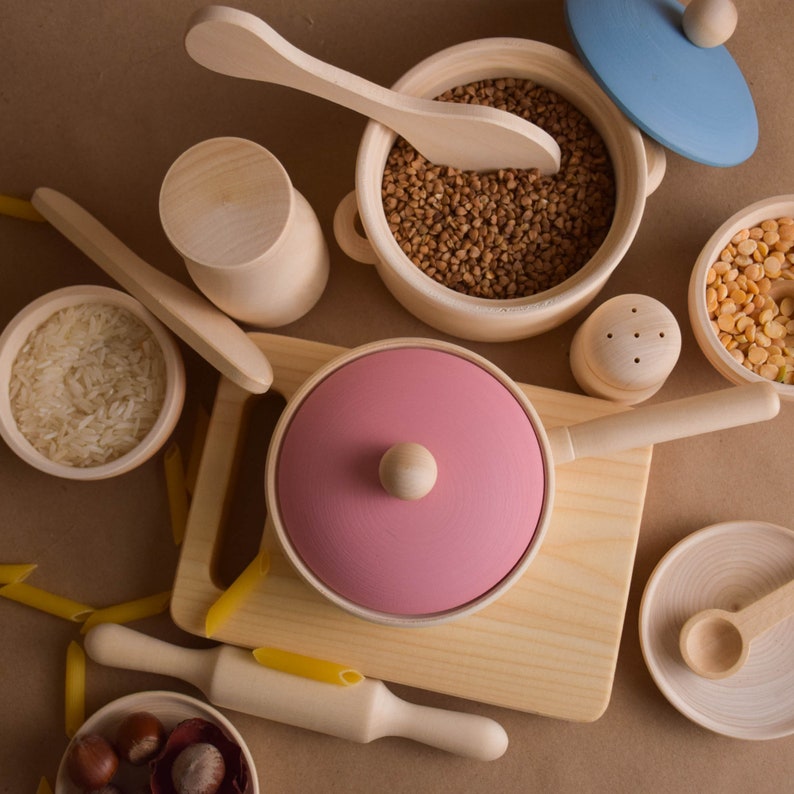 May include: A set of wooden play kitchen accessories, including a pink pot with a lid, a wooden spoon, a bowl of rice, a bowl of brown grains, a small wooden salt shaker, a bowl of yellow grains, and a wooden rolling pin.