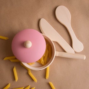 May include: A pink wooden lidded pot with a wooden handle and a wooden spoon. The pot is filled with yellow pasta and the spoon is resting on the pot. The pot is sitting on a brown surface.