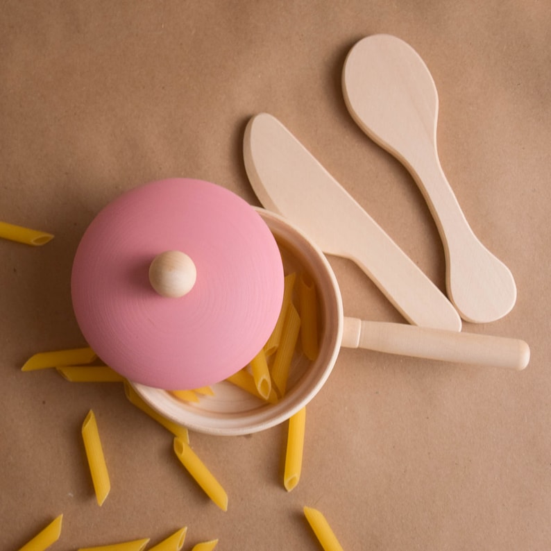 May include: A pink wooden lidded pot with a wooden handle and a wooden spoon. The pot is filled with yellow pasta and the spoon is resting on the pot. The pot is sitting on a brown surface.