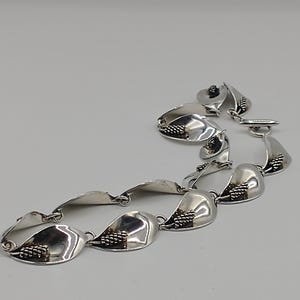 May include: A silver bracelet with a leaf-shaped design. Each leaf has a textured cluster of small, round details, possibly grapes. The bracelet is linked with small rings and a clasp. The design is reminiscent of a vine.