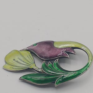 以下が含まれることがあります： スタイリッシュなデザインのフローラルブローチ。ブローチは、紫と黄色の花と緑の葉、銀色の金属の輪郭が特徴です。花の中心には小さな緑色の宝石があります。ブローチは装飾的なアクセサリーです。