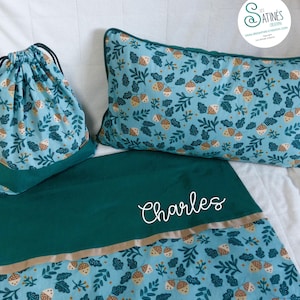 Peut inclure: Un ensemble de literie bleu sarcelle et vert comprenant un sac à cordon, un oreiller et une couverture. L'ensemble présente un motif répété de glands et de feuilles. La couverture est personnalisée avec le nom "Charles" en écriture blanche.