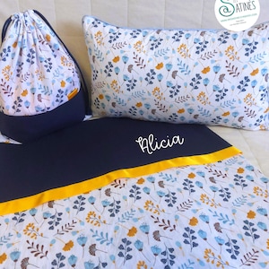 Può includere: Un set di biancheria da letto con un motivo floreale blu, giallo e marrone su sfondo bianco. Il set include un sacco con coulisse, un cuscino e una coperta con il nome "Alicia". La coperta ha un bordo di nastro giallo.