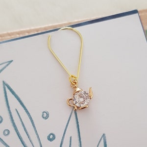 Puede incluir: Un charm de tetera de oro con una piedra de cristal transparente. El charm cuelga de un bucle de oro.