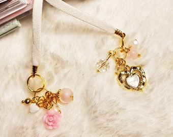 Könnte beinhalten: Weißes Band Lesezeichen mit goldenen Charms und rosa Perlen. Die Charms beinhalten ein Herz, eine Rose und eine kleine weiße Perle. Das Lesezeichen ist an einem schwarz-weißen Planer befestigt.