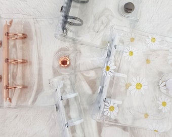 May include: Four clear plastic disc bound notebooks. One notebook has a daisy pattern, one has a silver ring binder, one has a gold ring binder, and one has a white ring binder.