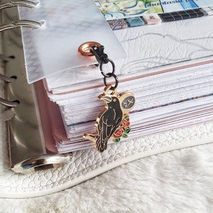 Könnte beinhalten: Ein goldfarbener Raben-Anhänger mit schwarzem Emaille und roten und weißen Emaille-Blumen. Der Anhänger ist an einer schwarzen Kette und einem goldfarbenen Ring befestigt.