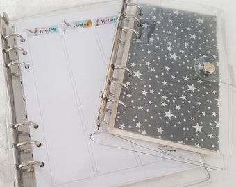 May include: Two clear plastic binders with silver rings. The top binder has a grey and white star pattern insert. The bottom binder has a white page with the words "Monday", "Tuesday", and "Wednesday" written on it.