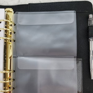 May include: A black leather planner with gold rings and two clear plastic pockets. The pockets are designed to hold cards, notes, or other small items.
