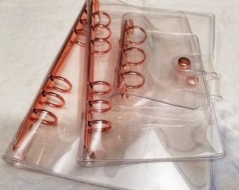 May include: Three clear plastic binders with rose gold rings and closures. The binders are stacked on top of each other.