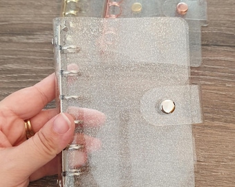 May include: Three clear plastic binders with silver rings and a sparkly finish. The binders are closed and stacked on top of each other. The top binder has a silver button closure.