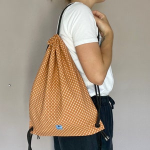 May include: A drawstring backpack with a repeating white floral pattern on a burnt orange background. The bag has black rope straps and a small blue label with a white graphic. The bag is worn over the shoulder.