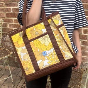 May include: A floral tote bag with a yellow and white flower pattern and brown trim. The bag has brown handles and a quilted base. A small blue tag is visible on the front of the bag.